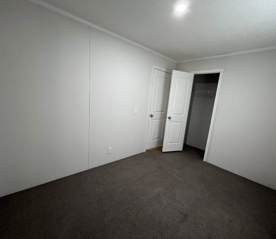 Empty room with white walls, dark carpet, open closet, and open door. A single ceiling light is turned on, and there is no furniture or decor visible.