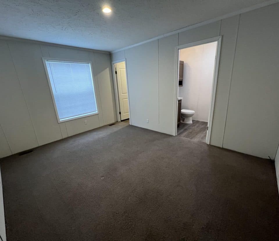 Empty carpeted room with a window on the left wall, a door to a small room, and an open doorway leading to a bathroom with visible toilet and vanity. Neutral walls and ceiling with a recessed light.