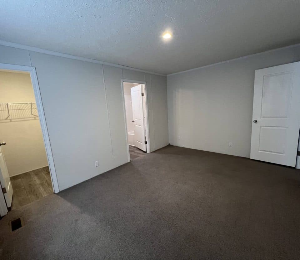 A carpeted bedroom with three white doors, one leading to a walk-in closet with shelves, another to a hallway, and a third closed. The room has white walls and a ceiling light.