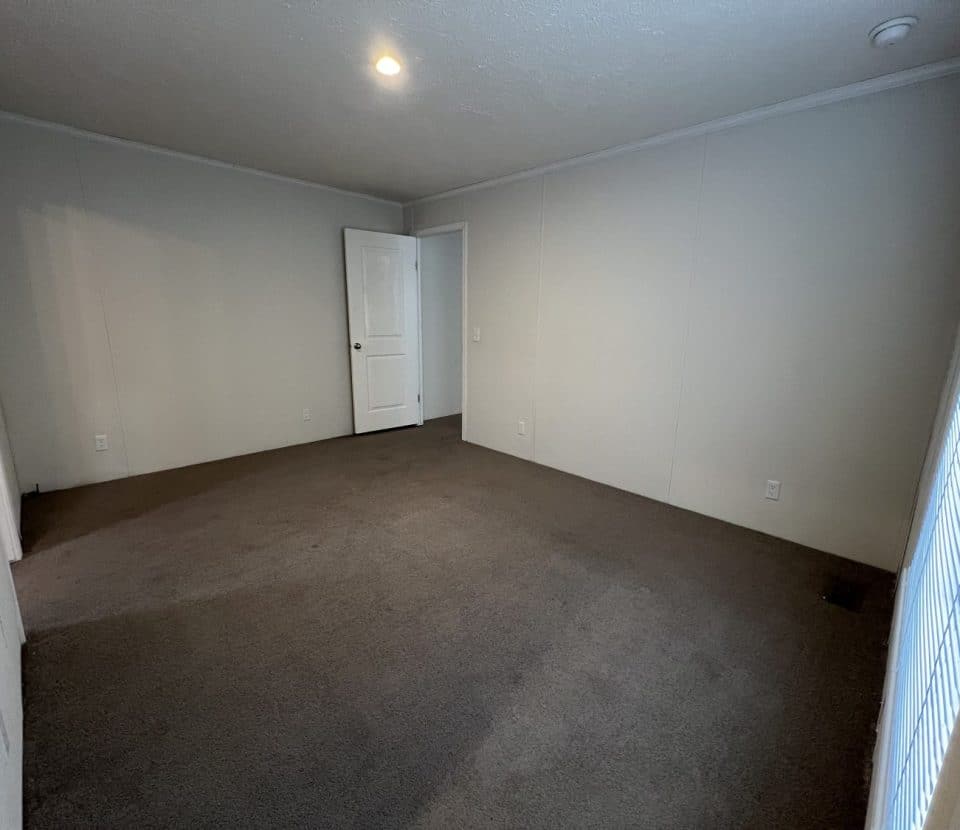 Empty room with beige walls, brown carpet, a single ceiling light, an open white door, and window blinds letting in daylight from the right side.