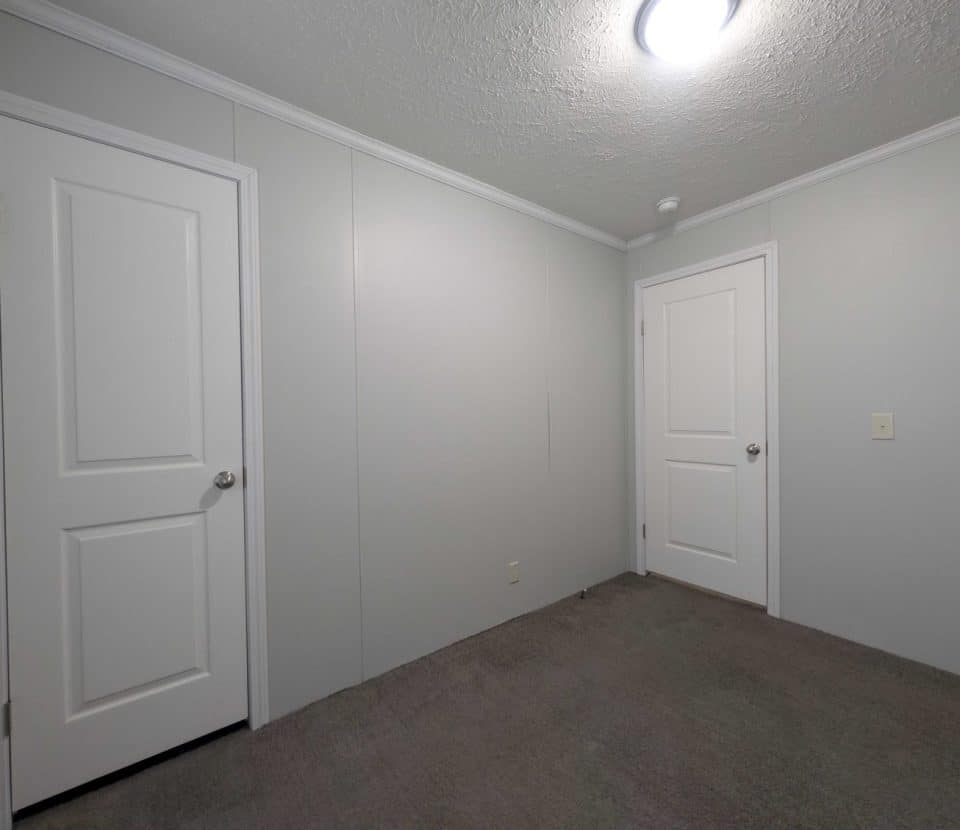 Empty room with light gray walls, two white doors, gray carpet, and a ceiling light. The room appears clean and simple with minimal decor and no furniture.