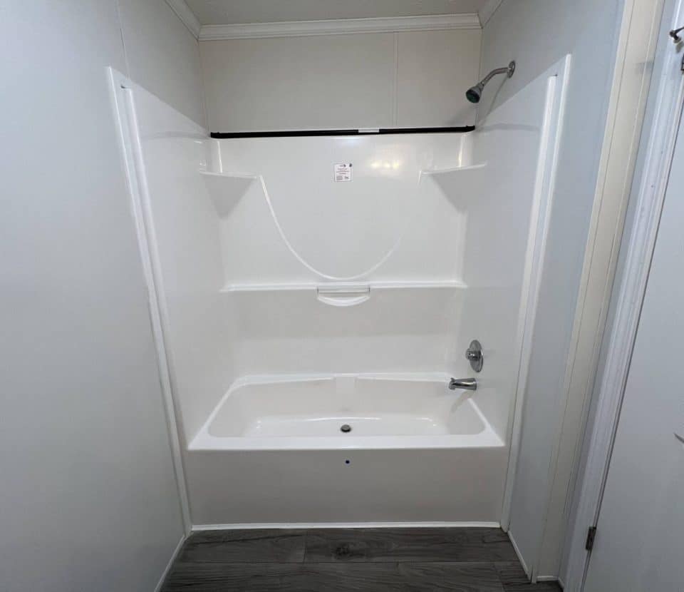 White bathtub and shower combo with a wall-mounted showerhead and built-in shelves, set against white walls and a wood-look floor. The photo is taken facing the tub directly.