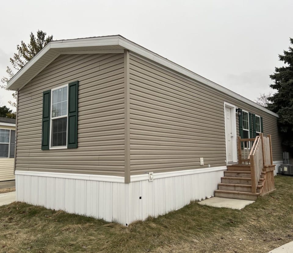 A beige manufactured home with green shutters and a small wooden staircase leading to the side door. The home sits on a grassy lot with a sidewalk and is next to another similar home.
