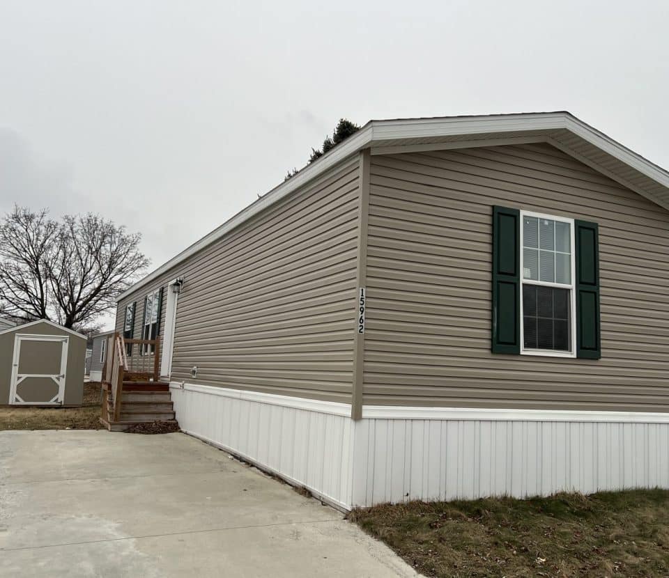 A beige mobile home with green shutters and white trim sits on a concrete driveway. Steps lead to the front door. Another similar structure and leafless trees are visible in the background under a cloudy sky.