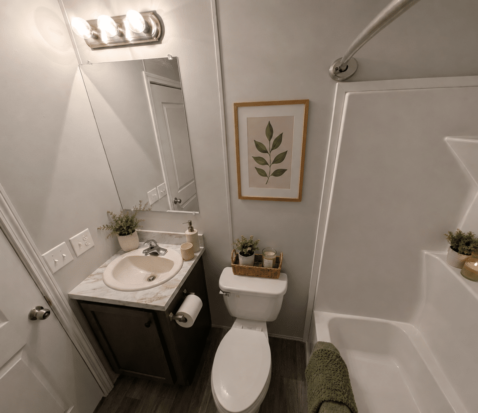 Small modern bathroom with a sink, mirror, toilet, and bathtub. Green accents include a framed leaf print, small potted plants, and a folded towel on the tub. Neutral colors and clean design.