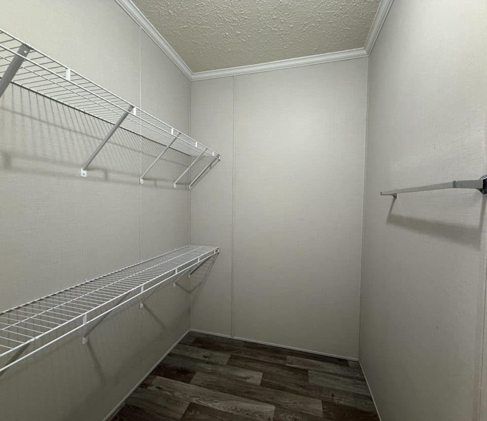 Empty walk-in closet with beige walls, crown molding, wood-style flooring, two white wire shelves on the left, and a metal bar for hanging clothes on the right wall.