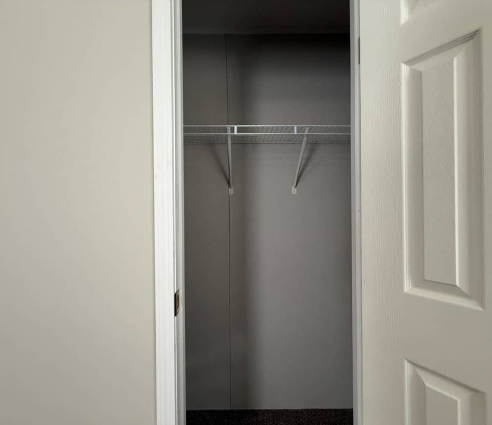 An empty closet with white walls, a white wire shelf, and an open white door. The closet floor is dark, and the space is well-lit.
