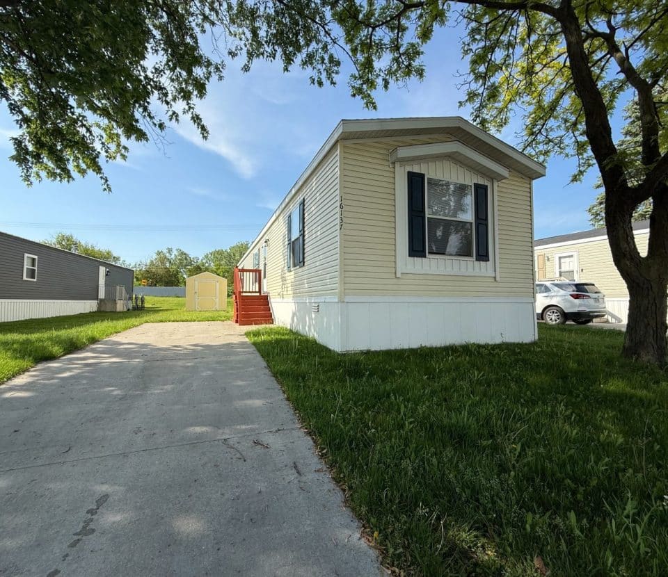 A beige mobile home with black shutters sits on a grassy lot with a concrete driveway, a red porch, and a shed in the backyard. Trees provide shade, and a neighboring home and parked car are visible.