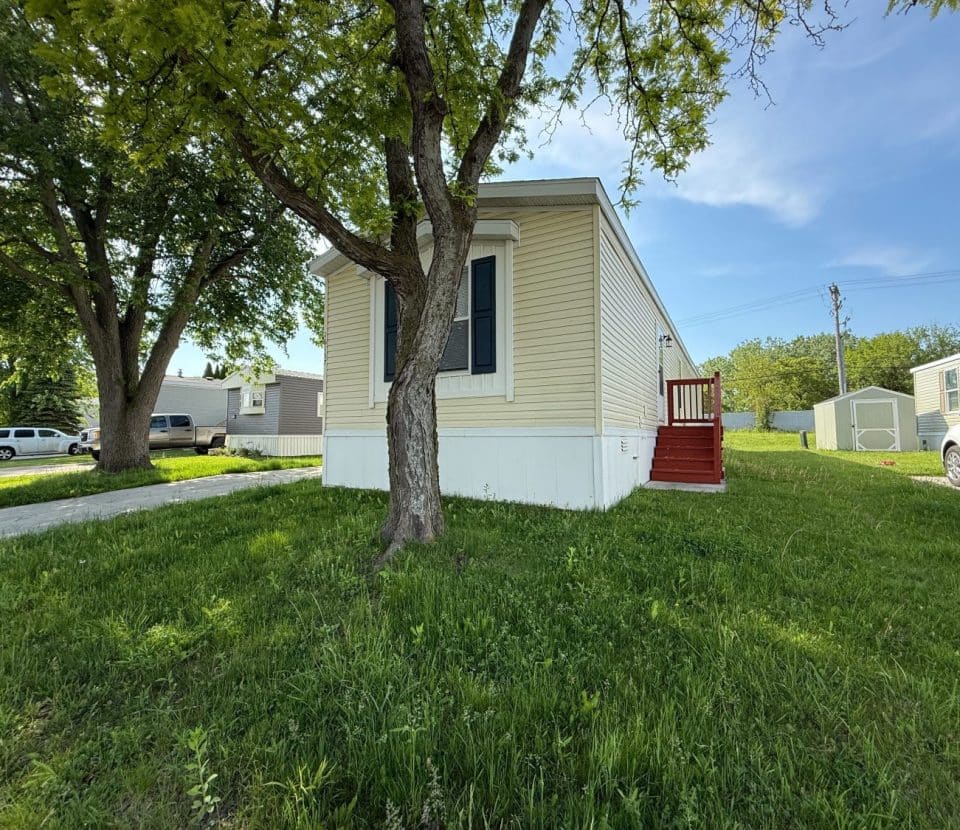 A beige mobile home with white trim and red steps sits on a grassy lot, shaded by a large tree. Nearby are other mobile homes, a white car, and a clear blue sky.