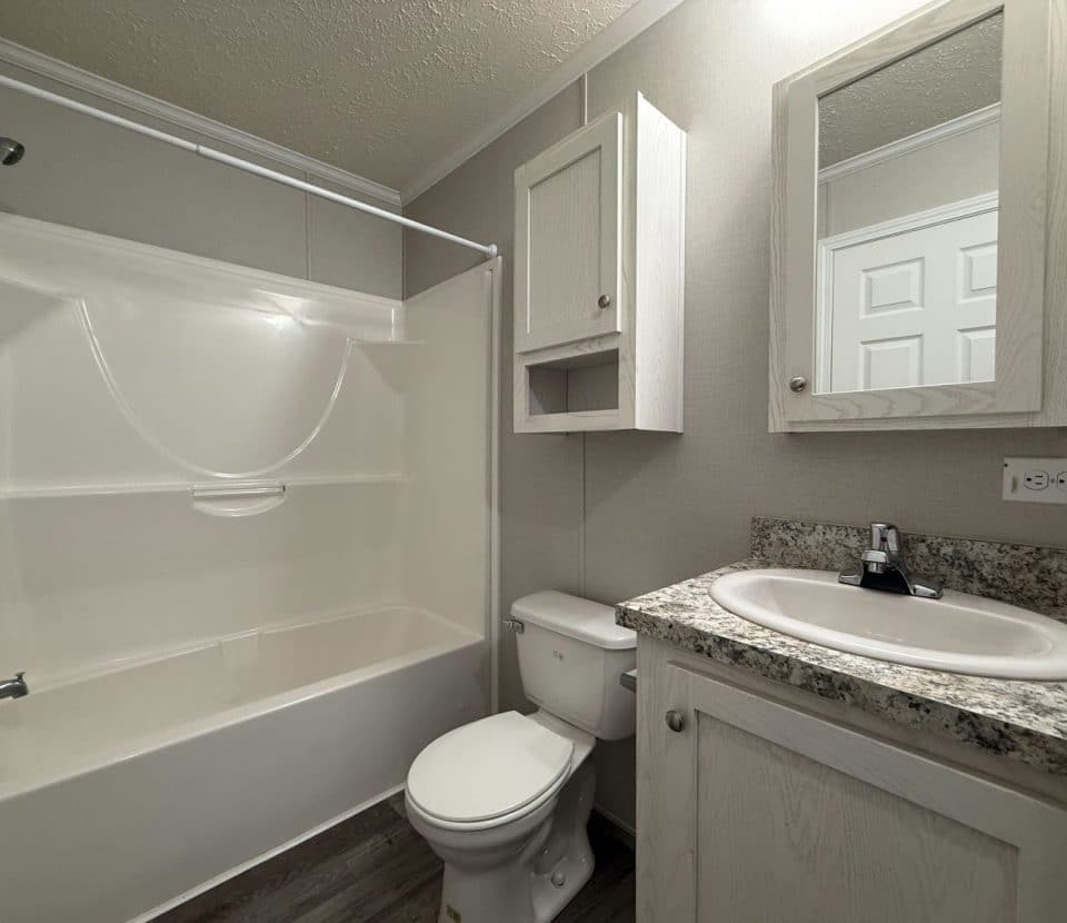 A small bathroom with a white bathtub-shower combo, toilet, and sink with a granite countertop. Light wood cabinets and a wall-mounted mirror are above the sink. The floor has wood-style vinyl planks.
