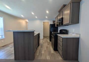 Modern kitchen with dark wood cabinets, marble countertops, gas stove, black refrigerator, microwave, and light wood flooring. Recessed ceiling lights brighten the space. A door and window are visible in the background.