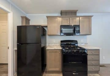A modern kitchen features a black refrigerator, a black gas stove with oven, and a black microwave. Light wood cabinets and marble-style countertops provide contrast. The space is clean and neatly organized.