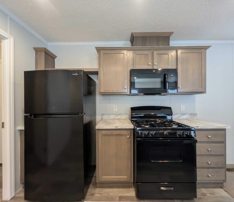 A modern kitchen features a black refrigerator, a black gas stove with oven, and a black microwave. Light wood cabinets and marble-style countertops provide contrast. The space is clean and neatly organized.