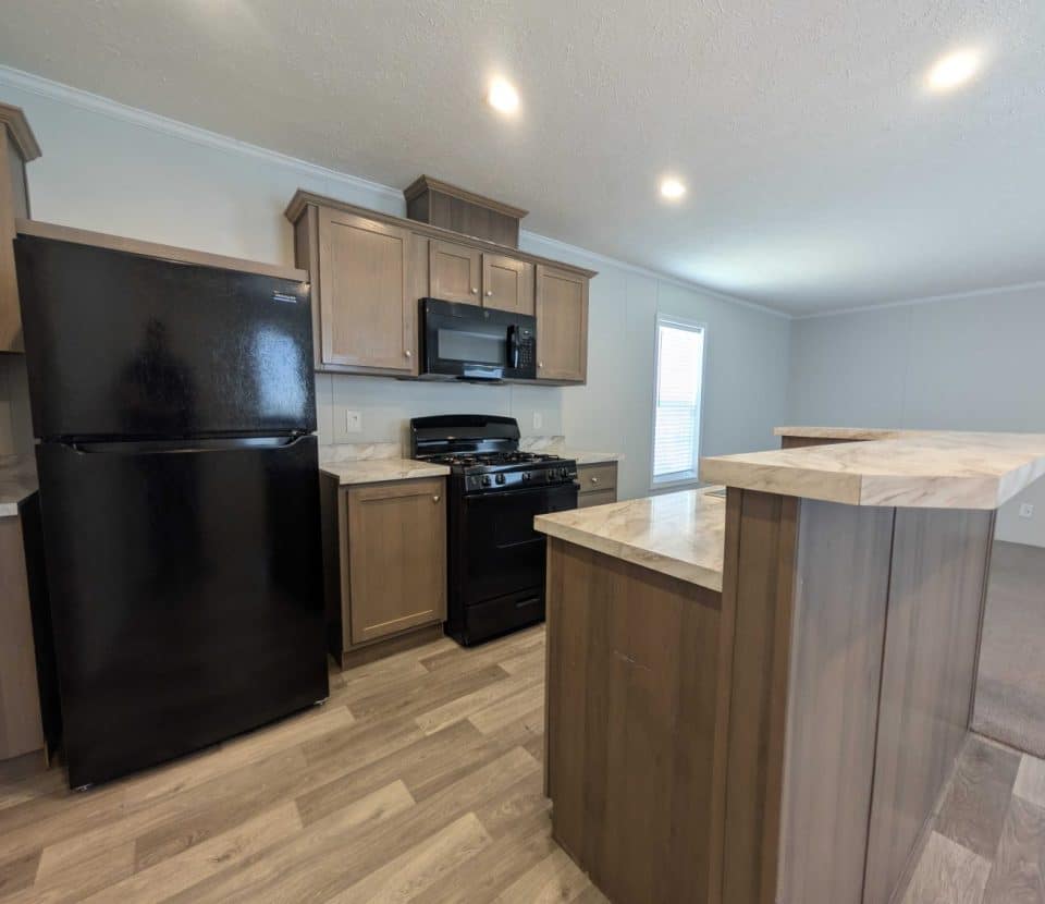 Modern kitchen with wood cabinets, black refrigerator, black stove and microwave, marble-patterned countertops, and light wood flooring. There is a bar-height counter and an adjacent open living area with carpet.