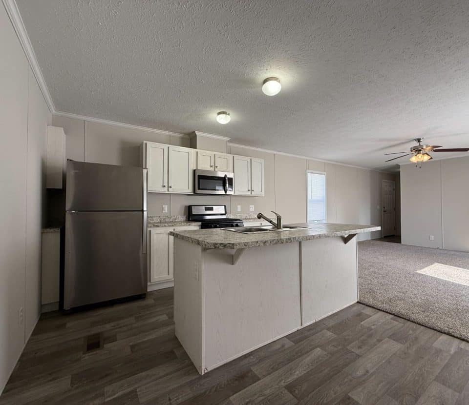 A modern kitchen with stainless steel appliances, white cabinets, and an island, opens to a carpeted living area with a ceiling fan and natural light from windows.