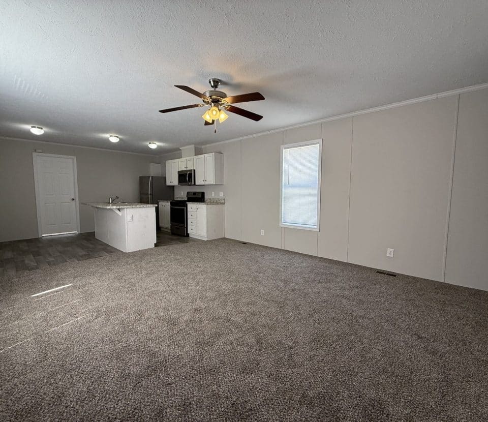 Empty living room with gray carpet, beige walls, a ceiling fan with lights, and an open kitchen area featuring white cabinets, stainless steel appliances, and tile flooring. One window lets in natural light.