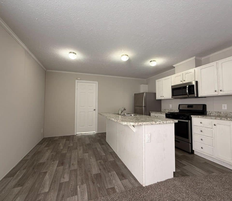 Modern kitchen with white cabinets, stainless steel appliances, granite countertops, and wood-look flooring. There is an island in the center and a white door in the background. The walls are beige, and ceiling lights are on.