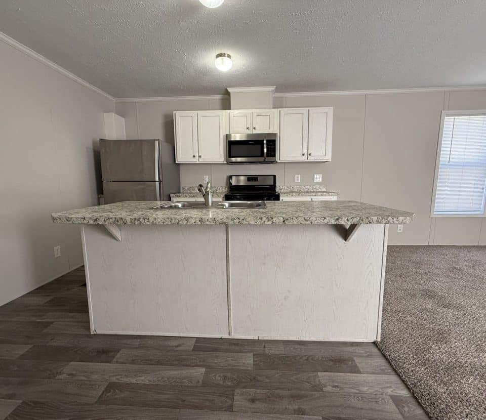 Modern kitchen with a marble countertop island, stainless steel refrigerator, microwave, stove, and white cabinets. The floor is hardwood, and there is carpeted flooring in the adjacent room.