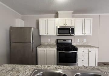Modern kitchen with white cabinets, stainless steel refrigerator, gas stove with oven, microwave, and a granite countertop with a double sink in the foreground.