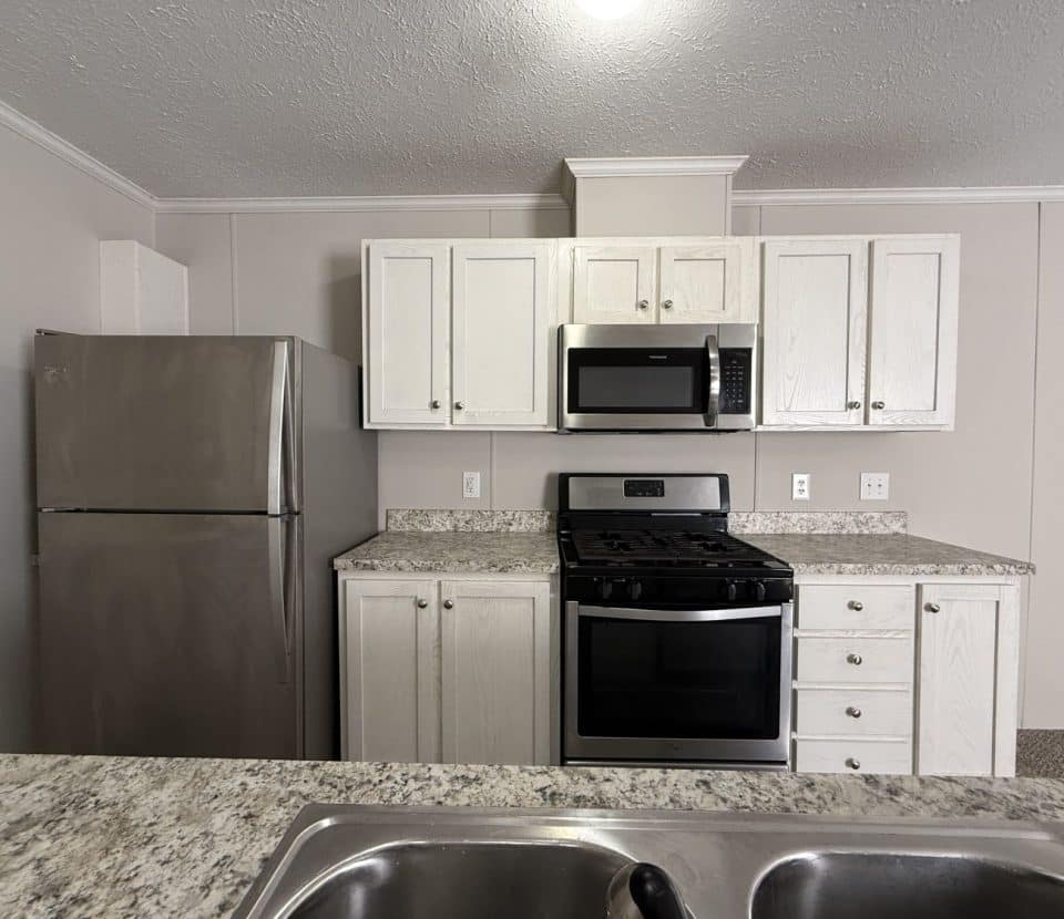 Modern kitchen with white cabinets, stainless steel refrigerator, gas stove with oven, microwave, and a granite countertop with a double sink in the foreground.