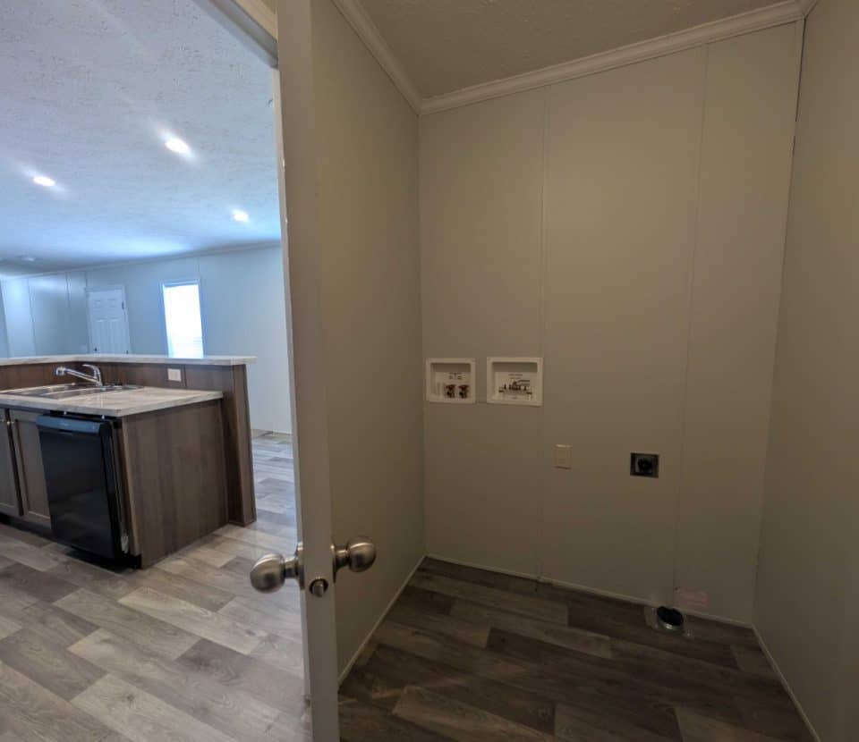 View from a doorway into a small laundry area with water hookups, next to an open kitchen with an island, dishwasher, and wood-style flooring throughout.