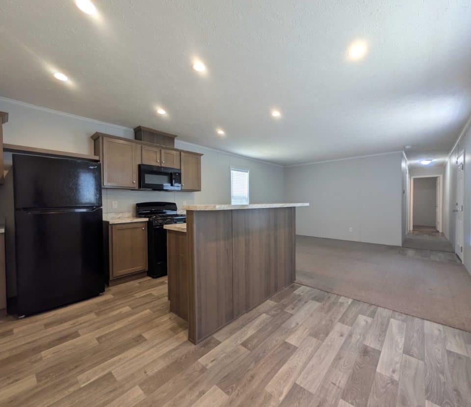 Modern kitchen with wood cabinets, black appliances, a center island, and wood-look flooring. The open layout leads to a carpeted living room and a hallway with natural light from windows.