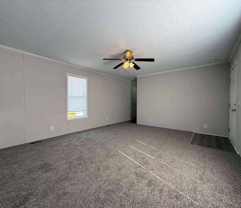 A spacious empty living room with gray carpet, beige walls, a ceiling fan with lights, and a window letting in natural light. Part of a kitchen counter is visible to the left.