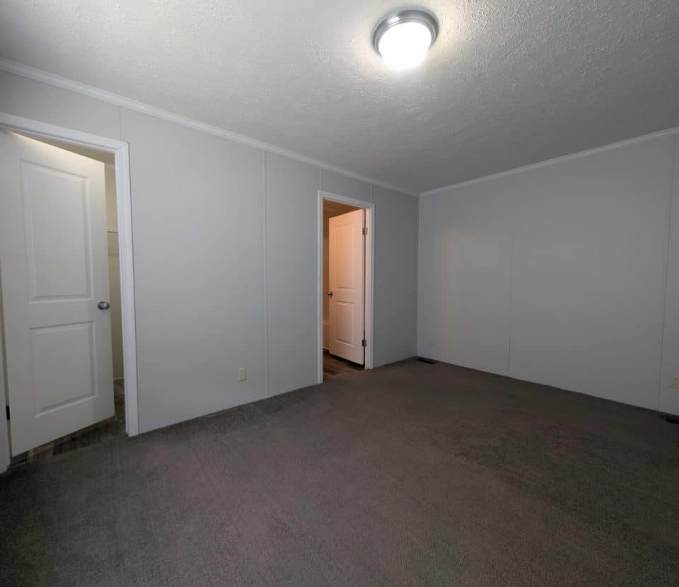 Empty room with gray carpet, white walls, ceiling light, and two open doors—one leading to a bright hallway, the other appears to lead to a bathroom.