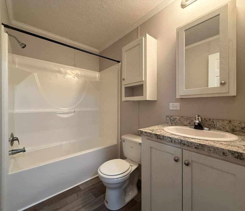 Small bathroom with a white bathtub-shower combo, toilet, wall cabinets, and a sink with a granite-patterned countertop. The floor is wood-style, and the walls are beige. Mirror and light fixture above the sink.
