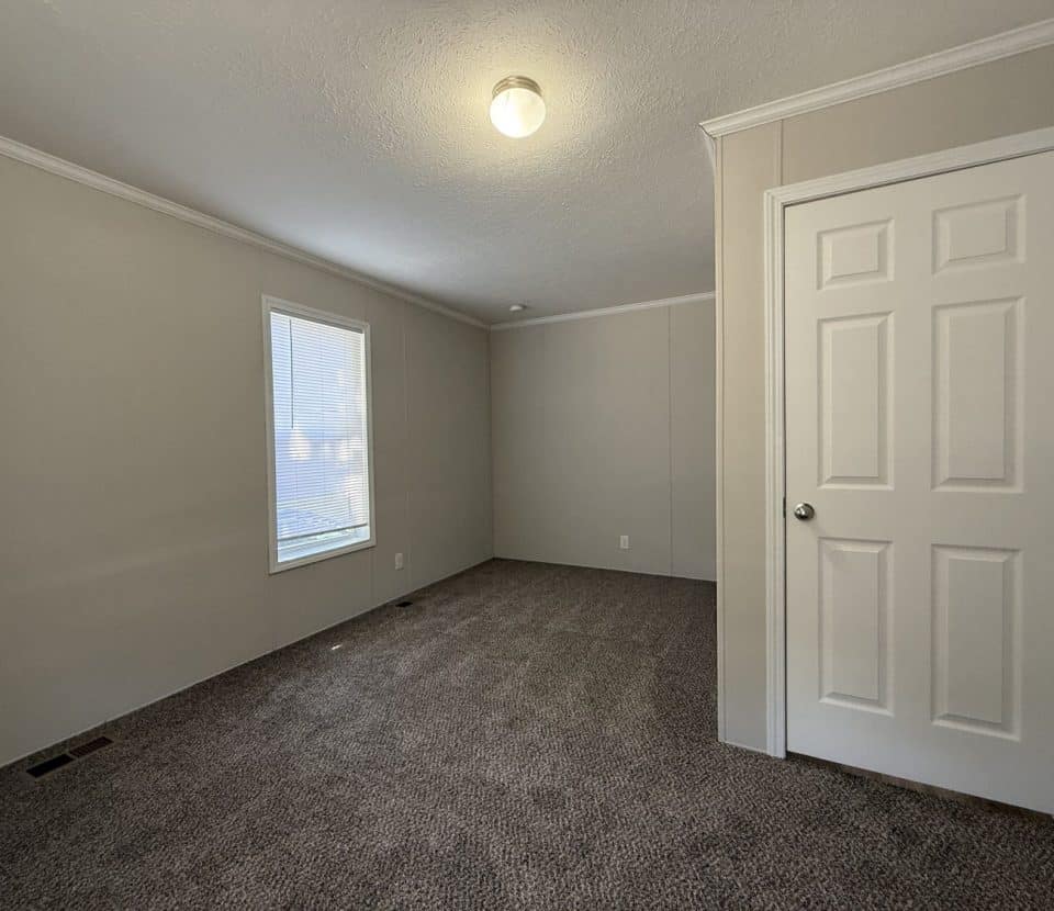 Empty room with beige walls, white trim, gray carpet, a single window with blinds, and a closed white door. A ceiling light fixture is turned on, illuminating the space.