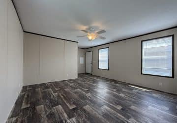 A spacious, empty room with light beige walls, dark trim, wood-style flooring, a white ceiling fan with lights, and large windows letting in natural light.