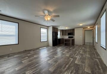 Spacious empty living area with wood-look flooring, beige walls, ceiling fan with light, large windows, and an open kitchen featuring dark cabinets, white countertops, and black appliances in the background.