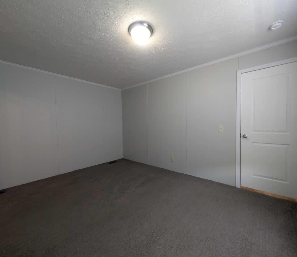 Empty room with gray carpet, light gray walls, a closed white door, and a ceiling light fixture. The space is undecorated and well-lit, with no furniture or windows visible.