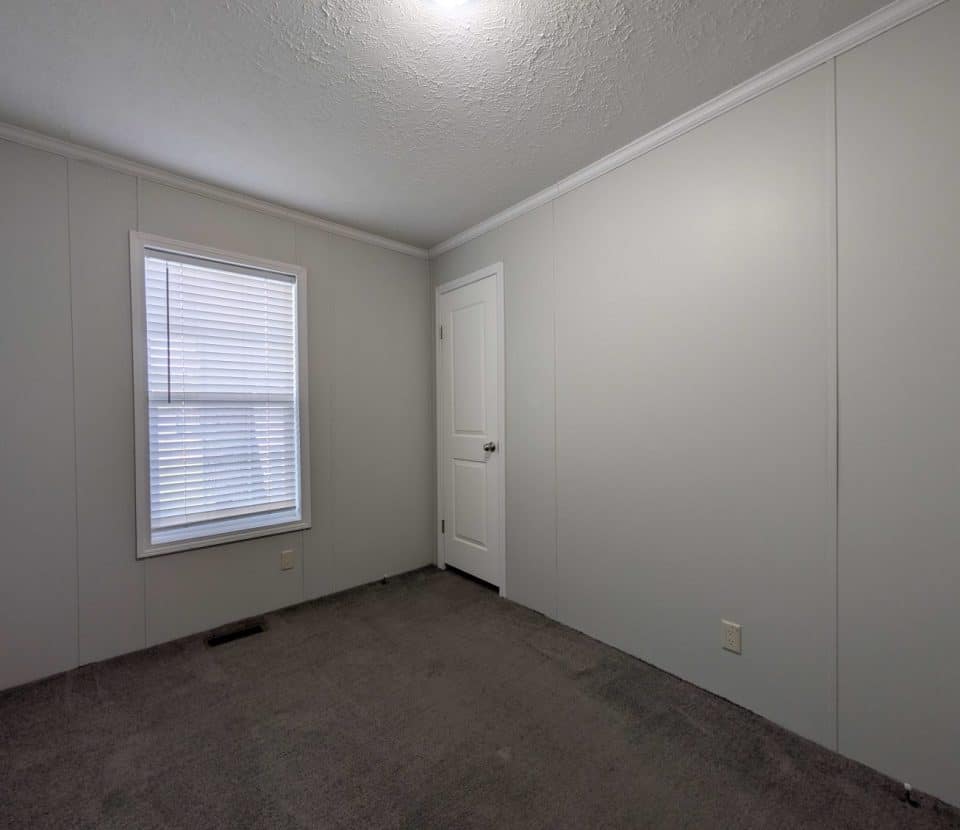 Empty room with gray carpet, light gray walls, a white door, and a single window with closed blinds. Ceiling is white with molding, and a light fixture is on. There are no furnishings or decorations.